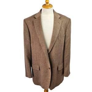 May include: A brown blazer with a two-button closure, notched lapels, and two front pockets. The blazer is displayed on a mannequin with a cream-colored shirt underneath. The fabric has a textured appearance.