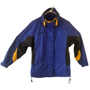 Könnte beinhalten: Eine blaue, schwarze und gelbe Columbia Jacke. Die Jacke hat einen hohen Kragen mit gelbem Besatz, schwarze Einsätze an den Seiten und Ärmeln sowie gelbe Akzente. Das Columbia-Logo ist auf der Brust sichtbar. Die Jacke hängt an einem weißen Kleiderbügel.