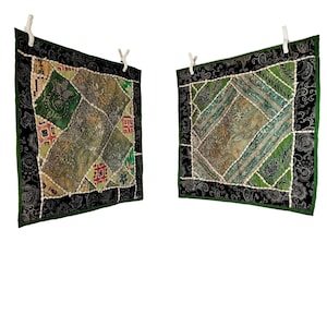 May include: Two square, patchwork textile wall hangings. Each features a central design of various green and tan fabrics, bordered by a black paisley pattern and a green edge. The textiles are suspended from white clothespins.