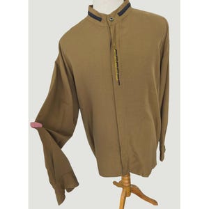 May include: A long-sleeved tan shirt featuring a mandarin collar with dark navy blue trim. The front placket displays a vertical row of small, rectangular yellow embroidered accents. The shirt has a button-up closure and long sleeves with cuffs.