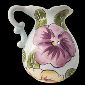 May include: A light blue ceramic pitcher with a floral design. The pitcher features a large purple pansy with green leaves and a smaller yellow pansy.