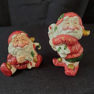 May include: A set of two ceramic Christmas elf salt and pepper shakers. The elf on the left is sitting down with a candy cane in his hand. The elf on the right is standing up and holding a bag of gifts. Both elves are wearing red and green clothing with gold accents.