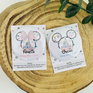 May include: Two round, white, Disney World themed buttons with a pink and blue watercolour design. The buttons feature a Mickey Mouse head silhouette with Cinderella's castle in the centre. The text on the buttons reads "Amelia Disney World 2024" and "Oscar Disney World 2024".