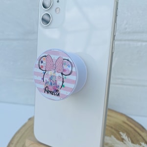 May include: A white phone case with a pink and white Minnie Mouse phone grip. The grip features a pink bow, glitter, and a pink and white striped background. The grip also has a picture of Cinderella's castle and the text "Amelia".