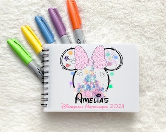 Puede incluir: Un libro de autógrafos blanco con espiral, con un lazo rosa y un dibujo del castillo de Cenicienta. El libro tiene el texto "Amelia's Disneyland Autographs 2024" impreso en la portada.