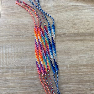 May include: A set of six colorful friendship bracelets with a chevron pattern. The bracelets are made with different colored threads and are tied together at the ends.