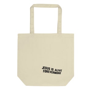 Op de afbeelding: Een beige tote bag met zwarte handvatten en de tekst "JESUS IS ALIVE FOREVERMORE" gedrukt op de voorkant.