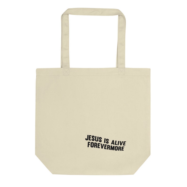 Op de afbeelding: Een beige tote bag met zwarte handvatten en de tekst "JESUS IS ALIVE FOREVERMORE" gedrukt op de voorkant.