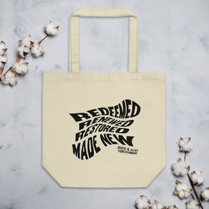Op de afbeelding: Een beige tote bag met zwarte tekst die "Redeemed, Renewed, Restored, Made New" en "Jesus is Alive Forevermore" zegt.