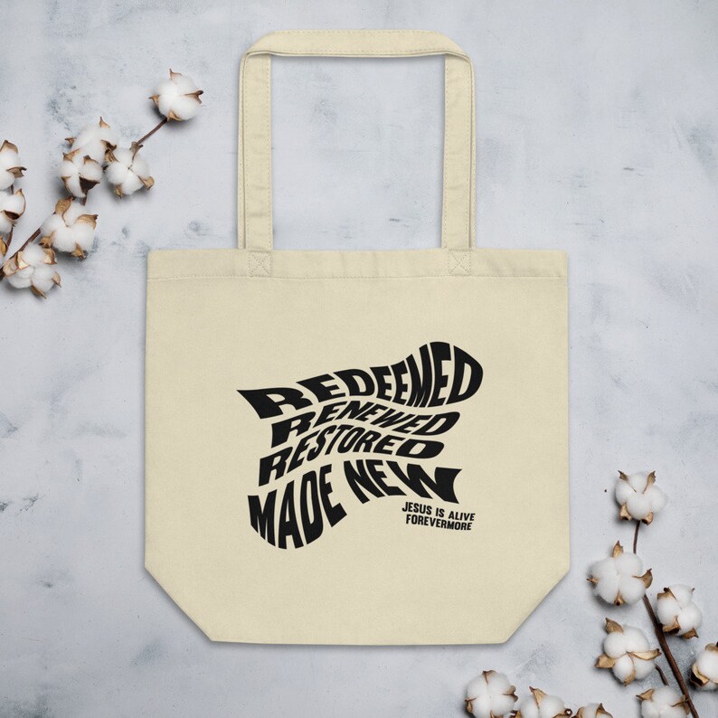 Op de afbeelding: Een beige tote bag met zwarte tekst die "Redeemed, Renewed, Restored, Made New" en "Jesus is Alive Forevermore" zegt.