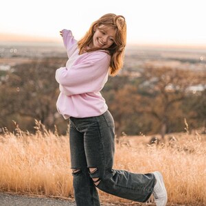 May include: A woman wearing a pink sweater and black jeans with ripped knees is smiling and posing with her arm outstretched. She is standing on a paved road with tall grass in the background.
