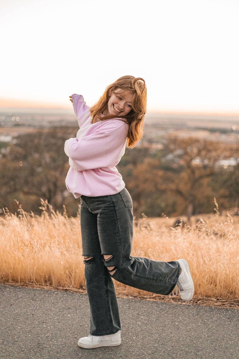 May include: A woman wearing a pink sweater and black jeans with ripped knees is smiling and posing with her arm outstretched. She is standing on a paved road with tall grass in the background.