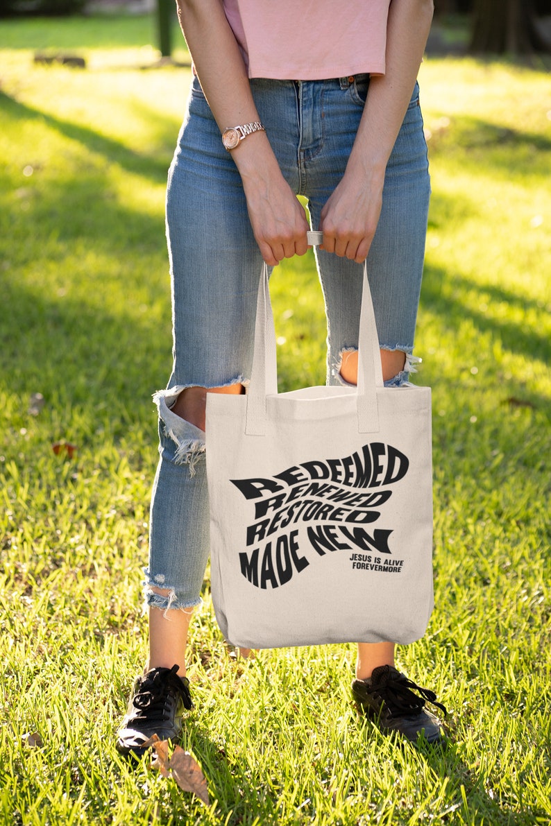 Op de afbeelding: Een witte tote bag met zwarte tekst die "Redeemed, Renewed, Restored, Made New" en "Jesus is Alive Forevermore" zegt.
