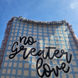 May include: A blue and white checkered woven throw blanket with the text "no greater love" in black cursive lettering.