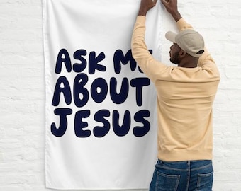 May include: A white banner with the text "ASK ME ABOUT JESUS" in large, bold, navy blue letters. A person is hanging the banner on a white brick wall.