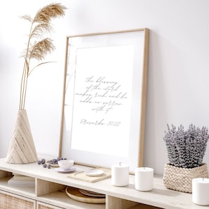 Puede incluir: Una impresión enmarcada con fondo blanco y texto negro que dice "La bendición del Señor enriquece, y no añade tristeza con ella. Proverbios 10:22". La impresión está colgada en una pared blanca sobre un estante de madera con velas, un cuenco de bayas y una planta en maceta.