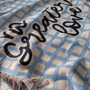 May include: A blue and white woven throw blanket with the text "The greater love" embroidered in black.