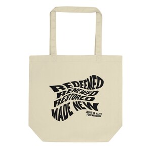 Op de afbeelding: Een beige tote bag met zwarte tekst die "Redeemed, Renewed, Restored, Made New" luidt in een draaiend ontwerp. De tekst "Jesus is Alive Forevermore" is onder het ontwerp gedrukt.