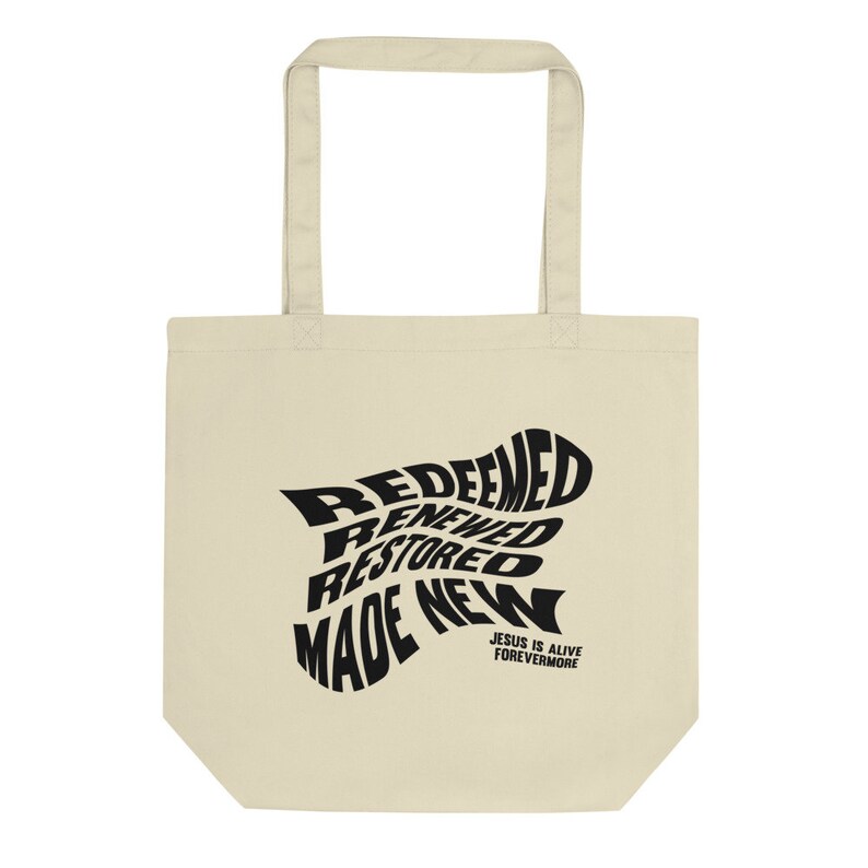 Op de afbeelding: Een beige tote bag met zwarte tekst die "Redeemed, Renewed, Restored, Made New" luidt in een draaiend ontwerp. De tekst "Jesus is Alive Forevermore" is onder het ontwerp gedrukt.