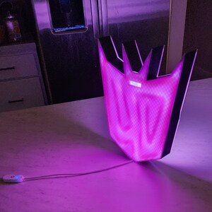 May include: A purple and black light-up sign shaped like the Decepticon symbol from the Transformers franchise. The sign is illuminated with a bright purple light.