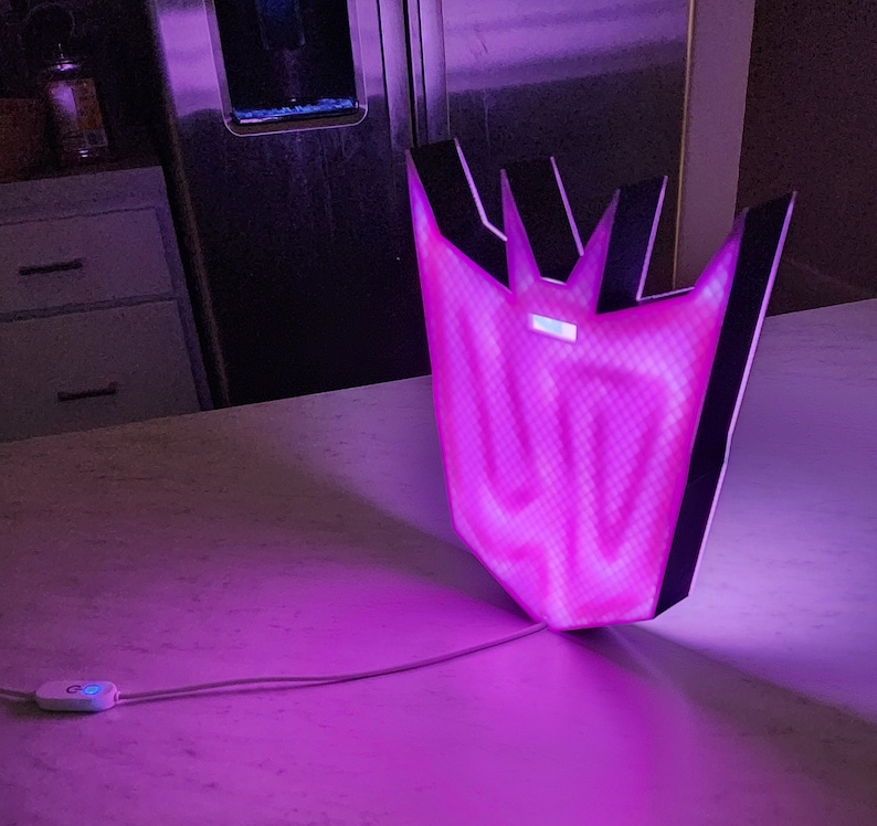 May include: A purple and black light-up sign shaped like the Decepticon symbol from the Transformers franchise. The sign is illuminated with a bright purple light.