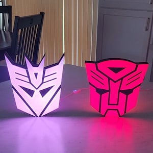 May include: Two Transformers logos, one in purple and one in red, are illuminated with LED lights. The logos are made of a hard plastic material and are shaped like the iconic Transformers symbols.