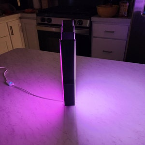 May include: A black, rectangular lamp with a pink light emanating from the sides. The lamp is sitting on a white countertop.