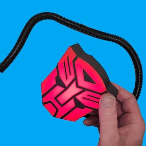 May include: A black and red plastic light-up toy shaped like the Autobot symbol from the Transformers franchise. The toy is attached to a black flexible tube.