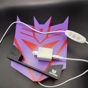 May include: A purple and red 3D printed Transformers logo with a black handle and a white power adapter and cord. The adapter has a white button with three symbols: a circle, a C, and a plus sign.