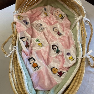 Puede incluir: Un moisés de mimbre con una manta acolchada rosa y verde con personajes de Peanuts bordados. La manta está doblada dentro del moisés.