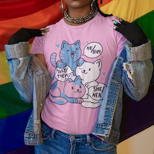 May include: A person wearing a pink t-shirt with a graphic of four cats and the text "He/Him", "They/Them", and "She/Her". The person is wearing a green turban, a denim jacket, and blue jeans. The person is standing in front of a rainbow flag.