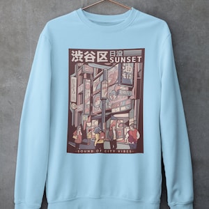 May include: Light blue sweatshirt with a graphic print of a city street scene at sunset. The graphic features a Japanese cityscape with neon signs and people walking. The text "渋谷区 SUNSET" is printed on the graphic. The text "SOUND OF CITY VIBES" is printed below the graphic.