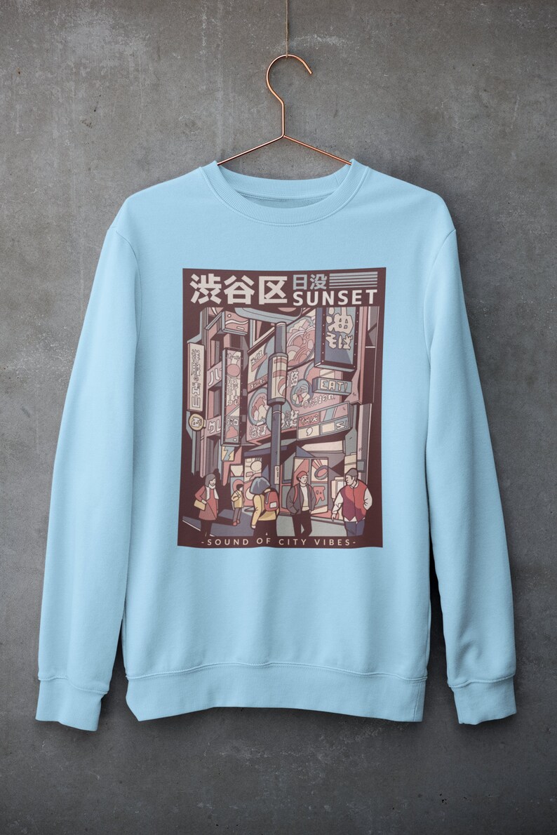 May include: Light blue sweatshirt with a graphic print of a city street scene at sunset. The graphic features a Japanese cityscape with neon signs and people walking. The text "渋谷区 SUNSET" is printed on the graphic. The text "SOUND OF CITY VIBES" is printed below the graphic.