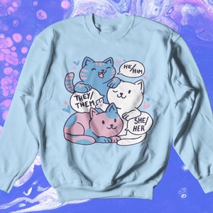 May include: A light blue sweatshirt with a graphic of three cartoon cats. The cats are stacked on top of each other. The top cat is blue and has a speech bubble that says "He/Him". The middle cat is white and has a speech bubble that says "She/Her". The bottom cat is pink and has a speech bubble that says "They/Them".