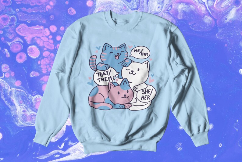 May include: A light blue sweatshirt with a graphic of three cartoon cats. The cats are stacked on top of each other. The top cat is blue and has a speech bubble that says "He/Him". The middle cat is white and has a speech bubble that says "She/Her". The bottom cat is pink and has a speech bubble that says "They/Them".