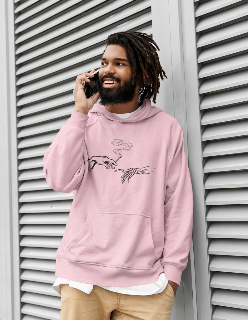 May include: A pink hoodie with a black and white graphic print of two hands, one human and one skeletal, passing a cigarette. The graphic is a parody of Michelangelo's The Creation of Adam.