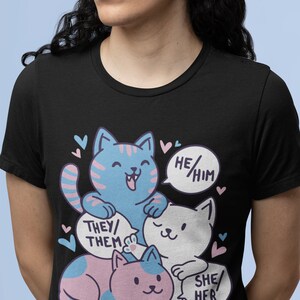 May include: Black t-shirt with a graphic of three cartoon cats. The cats are stacked on top of each other. The top cat is blue and says "He/Him". The middle cat is white and says "They/Them". The bottom cat is pink and says "She/Her".