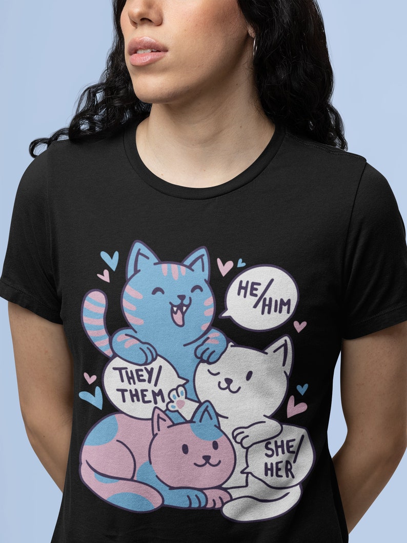 May include: Black t-shirt with a graphic of three cartoon cats. The cats are stacked on top of each other. The top cat is blue and says "He/Him". The middle cat is white and says "They/Them". The bottom cat is pink and says "She/Her".