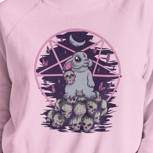 May include: A pink sweatshirt with a graphic of a gray bunny rabbit wearing a cross on its head. The bunny is sitting on a pile of skulls with a pentagram and crescent moon in the background.