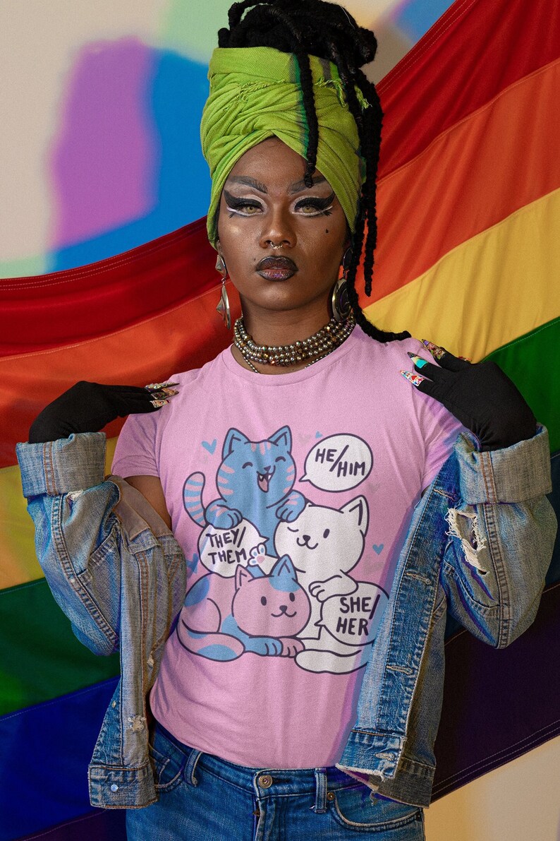 May include: A person wearing a pink t-shirt with a graphic of four cats and the text "He/Him", "They/Them", and "She/Her". The person is wearing a green turban, a denim jacket, and blue jeans. The person is standing in front of a rainbow flag.