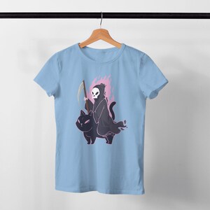 K&ouml;nnte beinhalten: Ein hellblaues T-Shirt mit einem Motiv des Sensenmanns, der auf einer schwarzen Katze reitet. Der Sensenmann h&auml;lt eine Sense und hat einen Sch&auml;del als Kopf. Die Katze ist von einer rosa Flamme umgeben.