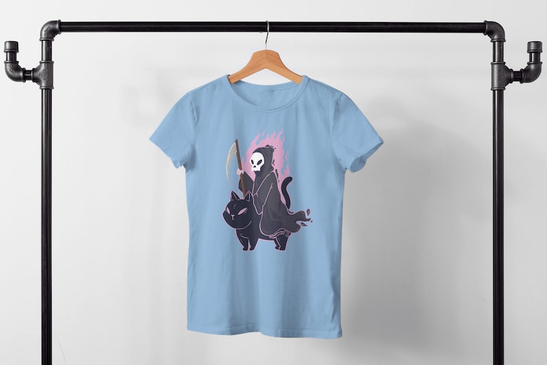 K&ouml;nnte beinhalten: Ein hellblaues T-Shirt mit einem Motiv des Sensenmanns, der auf einer schwarzen Katze reitet. Der Sensenmann h&auml;lt eine Sense und hat einen Sch&auml;del als Kopf. Die Katze ist von einer rosa Flamme umgeben.