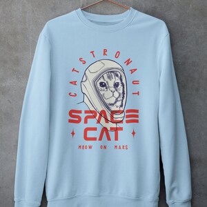 May include: Light blue sweatshirt with a graphic of a cat wearing an astronaut helmet. The text reads "Catstronaut Space Cat Meow On Mars".