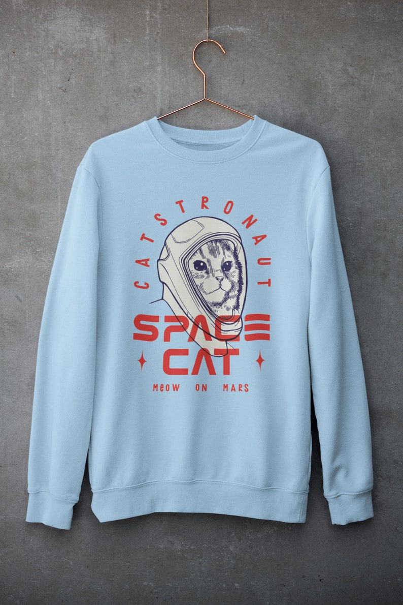 May include: Light blue sweatshirt with a graphic of a cat wearing an astronaut helmet. The text reads "Catstronaut Space Cat Meow On Mars".