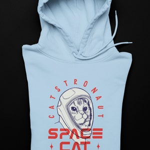 May include: A light blue hoodie with a graphic of a cat wearing an astronaut helmet. The text on the graphic reads "Catstronaut Space Cat Meow On Mars".