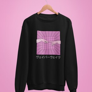 May include: Black sweatshirt featuring a pink and white graphic of two hands reaching towards each other within a 3D grid. Japanese text is below the graphic. The sweatshirt is hanging on a wooden hanger against a pink background.