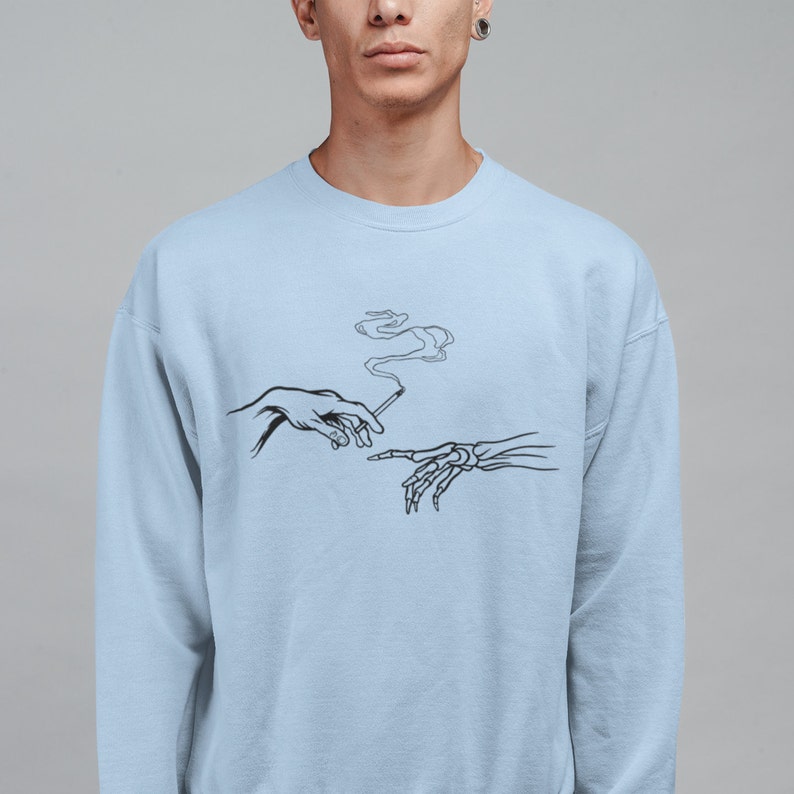May include: Light blue sweatshirt with a black and white graphic of two hands reaching towards each other, one hand is skeletal and the other is human. The skeletal hand is holding a cigarette with smoke rising from it.