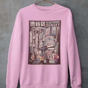 May include: Pink sweatshirt with a graphic print of a city street scene at sunset. The graphic features a Japanese cityscape with neon signs and people walking. The text "渋谷日没 SUNSET" is printed above the graphic. The text "SOUND OF CITY VIBES" is printed below the graphic.