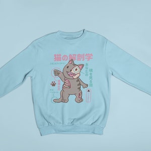 May include: A light blue sweatshirt featuring a cartoon cat anatomy illustration. The cat is labelled in Japanese with the text "猫の解剖学", which translates to "Cat Anatomy".