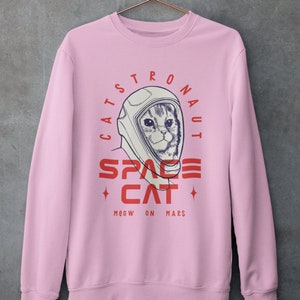 May include: Pink sweatshirt with a graphic of a cat wearing an astronaut helmet. The text reads "Catstronaut Space Cat Meow On Mars".